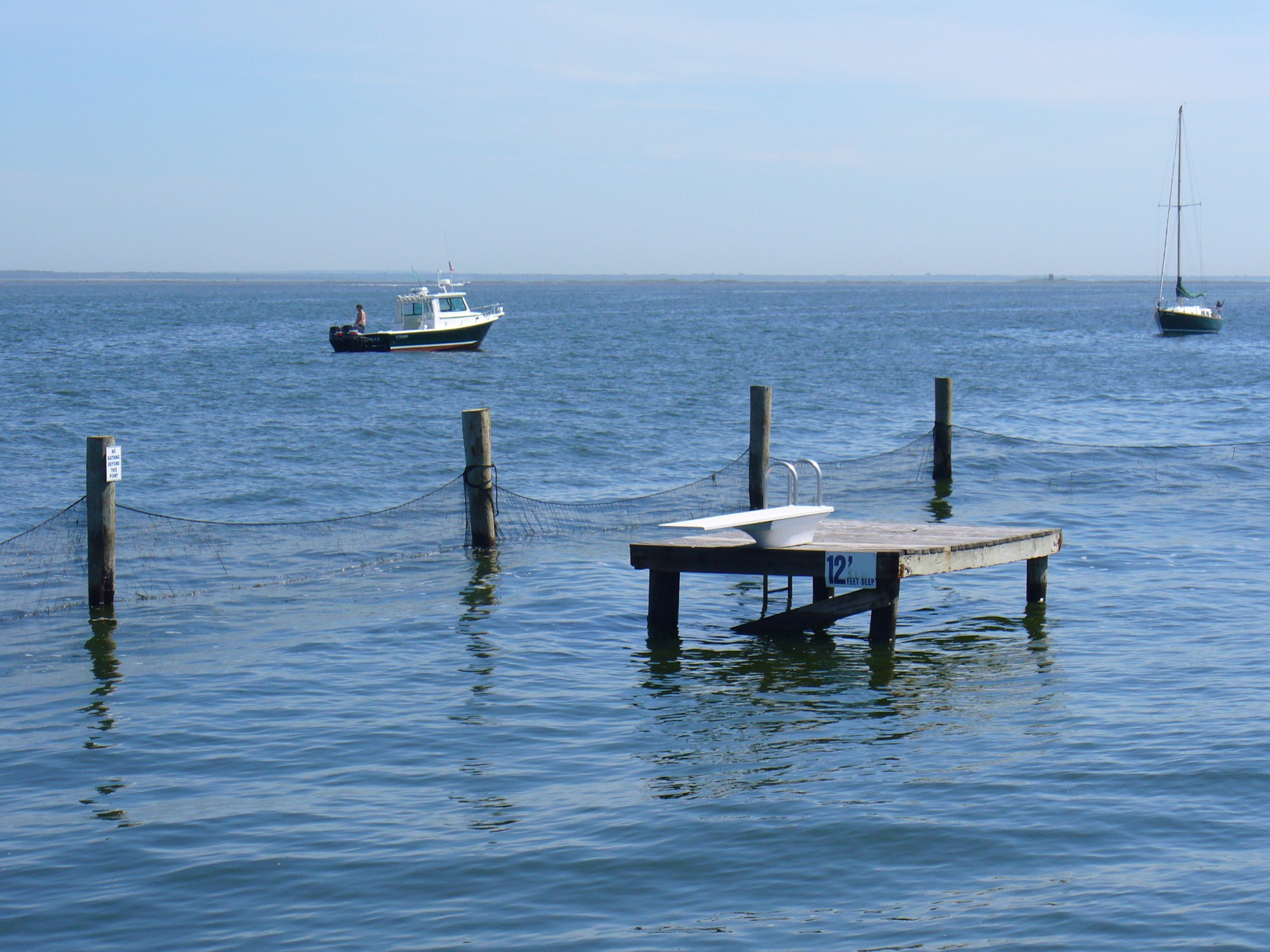 A small swimming dock with a diving board on the Great South Bay, a boat and a sailboat beyond