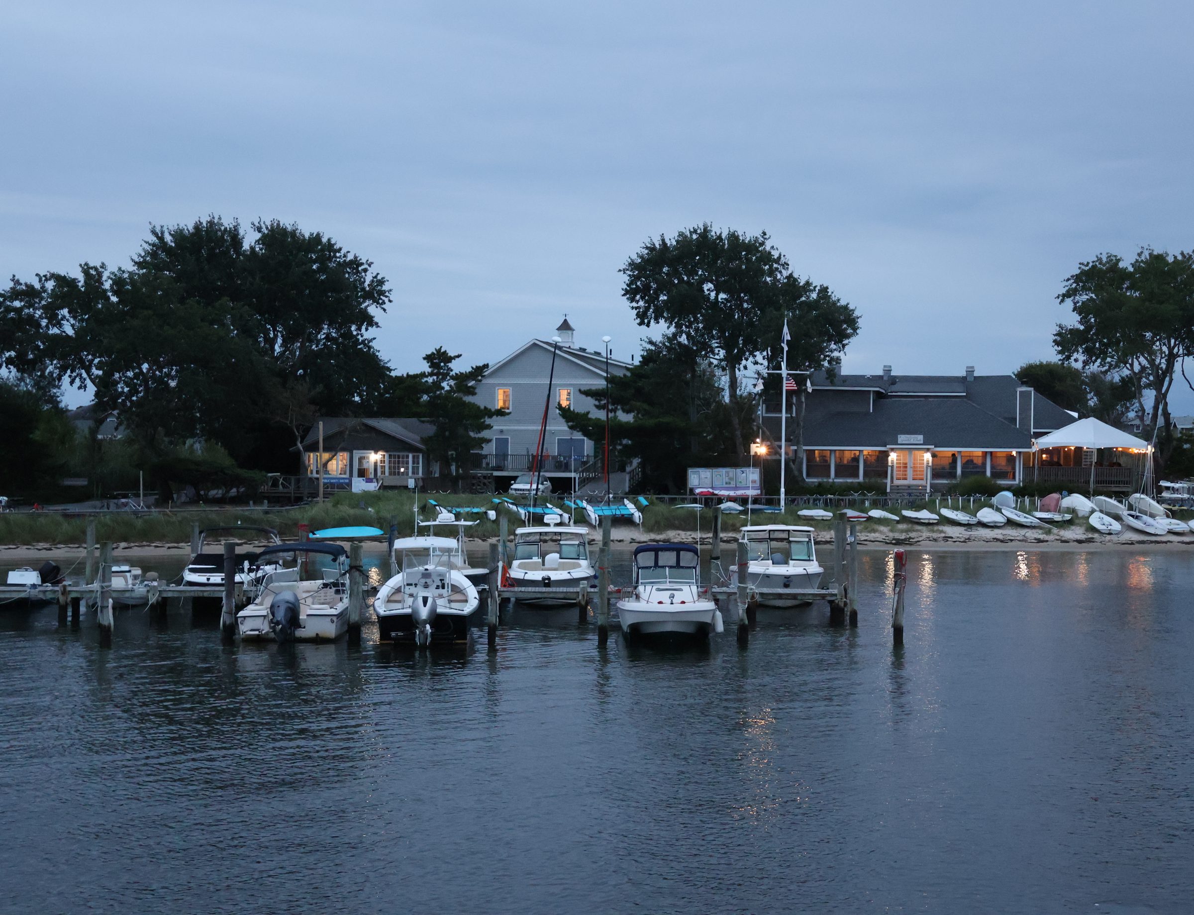 Saltaire harbor at dusk: boats at the docks, sailboats pulled up on the sand, the Yacht Club lit behind them