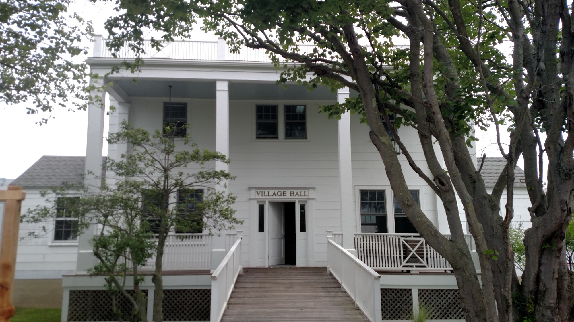The white clapboard Village Hall from the front: two-storey porch, boardwalk ramp to the door, trees in front